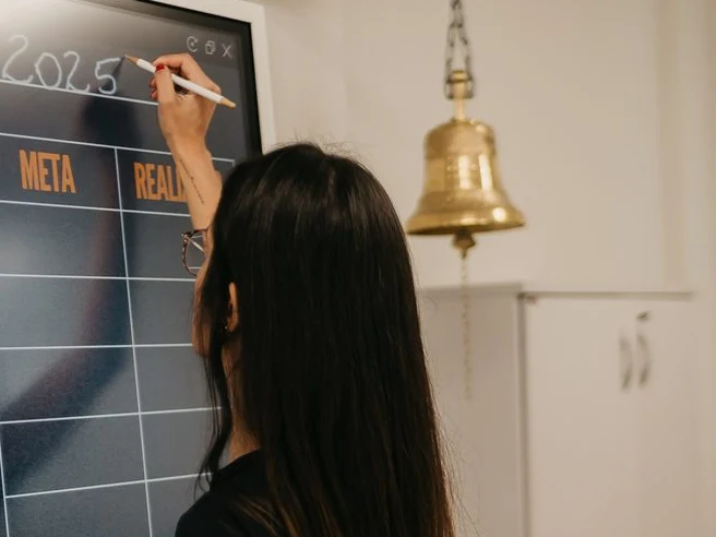 Pessoa de pé escrevendo o ano com caneta digital no alto de um painel de metas na parede, ao lado de um sino, com a grade de meta e realizado ainda vazia