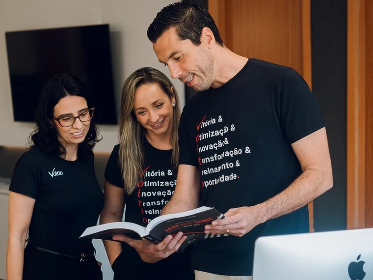 Thiago Coutinho em pé, de camiseta preta da Voitto, folheando um livro de Lean Seis Sigma aberto nas mãos, ao lado de duas colaboradoras também em pé que acompanham a leitura pela mesma página