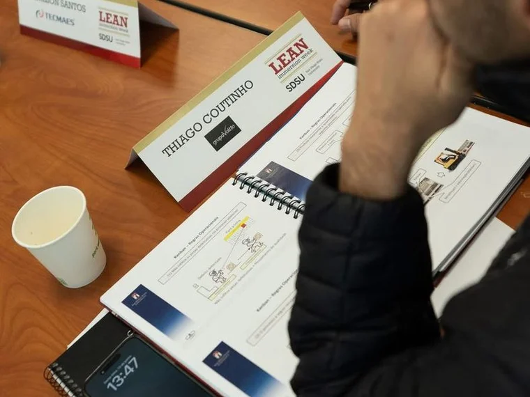 Braço e ombro de uma pessoa sentada a uma mesa de sala de aula, ao lado de uma placa de identificação, com uma apostila aberta na página de regras operacionais de kanban, um copo de papel e um caderno espiral sobre a mesa