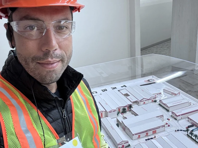 Thiago de capacete, óculos de proteção e colete refletivo em visita técnica, ao lado da maquete de uma planta industrial vista de cima