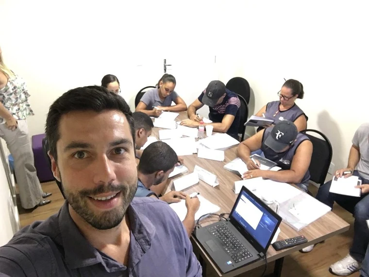 Thiago em primeiro plano num treinamento dentro da empresa, com participantes de boné e colete sentados ao redor de uma mesa preenchendo formulários impressos e um notebook aberto ao lado