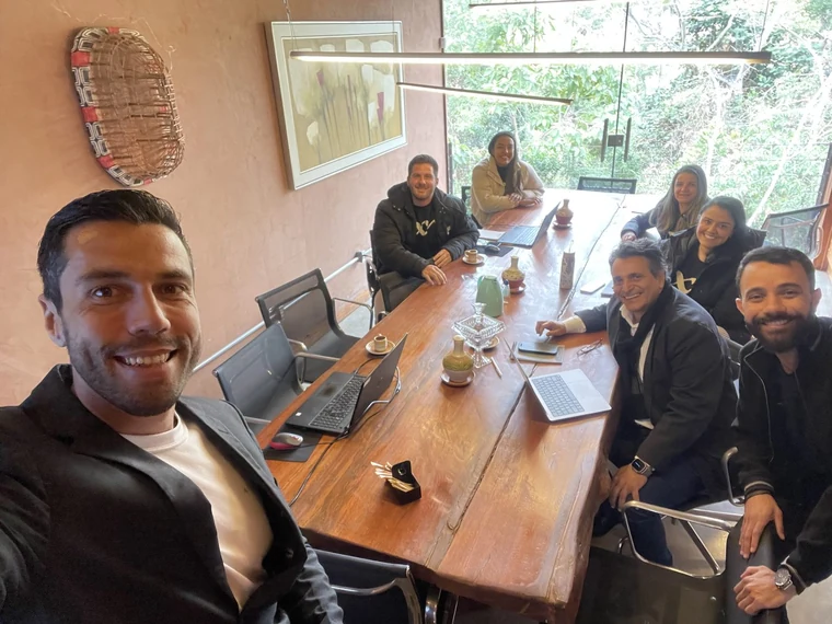 Thiago Coutinho em primeiro plano, de blazer sobre camiseta branca, com seis colegas sentados ao redor de uma mesa comprida de madeira maciça, notebooks e xícaras de café sobre a mesa e uma parede de vidro ao fundo dando para vegetação densa