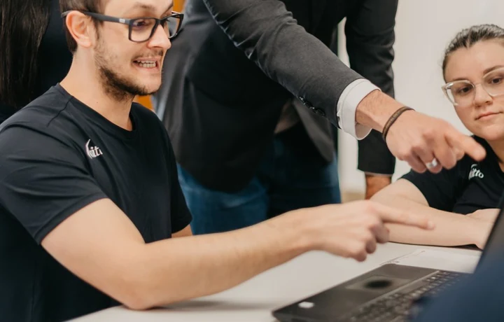 Profissional da Voitto de pé apontando a tela do notebook enquanto um colega sentado aponta o mesmo ponto, com uma terceira pessoa acompanhando