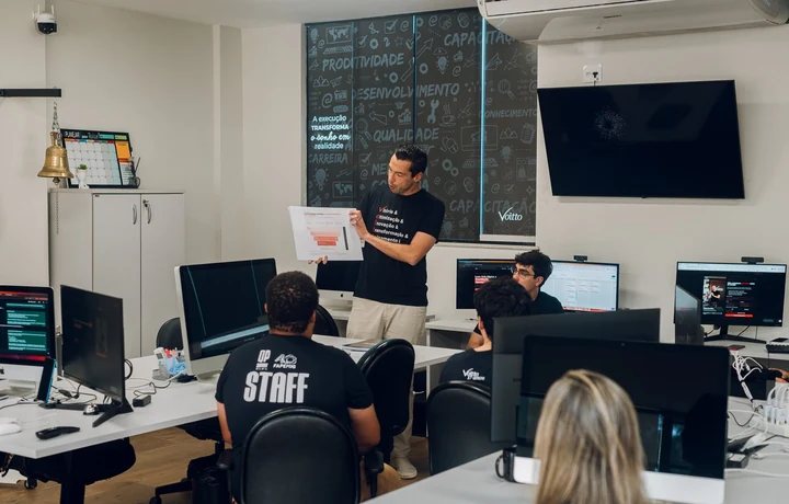 Apresentação em pé para a equipe sentada às mesas, com uma folha impressa na mão, em sala com computadores