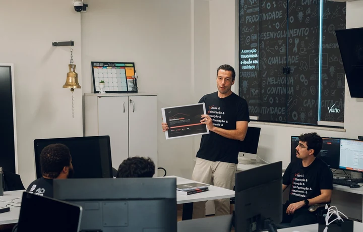 Homem em pé apresentando um quadro impresso a colegas sentados em estações de trabalho