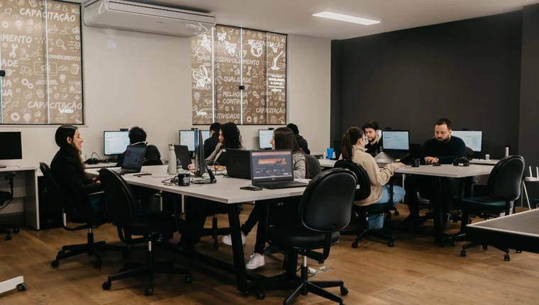 Equipe da Voitto trabalhando em computadores no escritório, com painéis de qualidade e melhoria contínua ao fundo