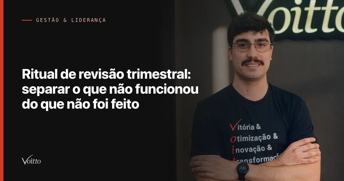 Colaborador da Voitto de camiseta preta e braços cruzados, sorrindo diante do letreiro Voitto no escritório, com o texto Ritual de revisão trimestral: separar o que não funcionou do que não foi feito sobre fundo escuro à esquerda
